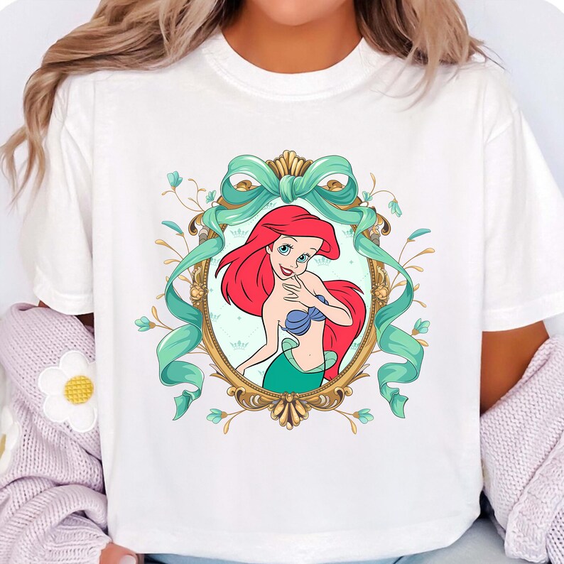 Op de afbeelding: Wit T-shirt met een cartoon illustratie van Ariel uit De Kleine Zeemeermin. Ariel heeft rood haar, een blauwe top en een groene staart, omlijst door een gouden ovaal en een turkoois lint. Bloemenaccenten omringen het frame.