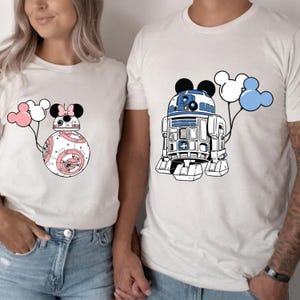 May include: Two white t-shirts with cartoon graphics. One shirt features a BB-8 droid design with a Minnie Mouse bow and pink balloons. The other shirt has an R2-D2 droid design with Mickey Mouse ears and white and blue balloons. The shirts are a light color.