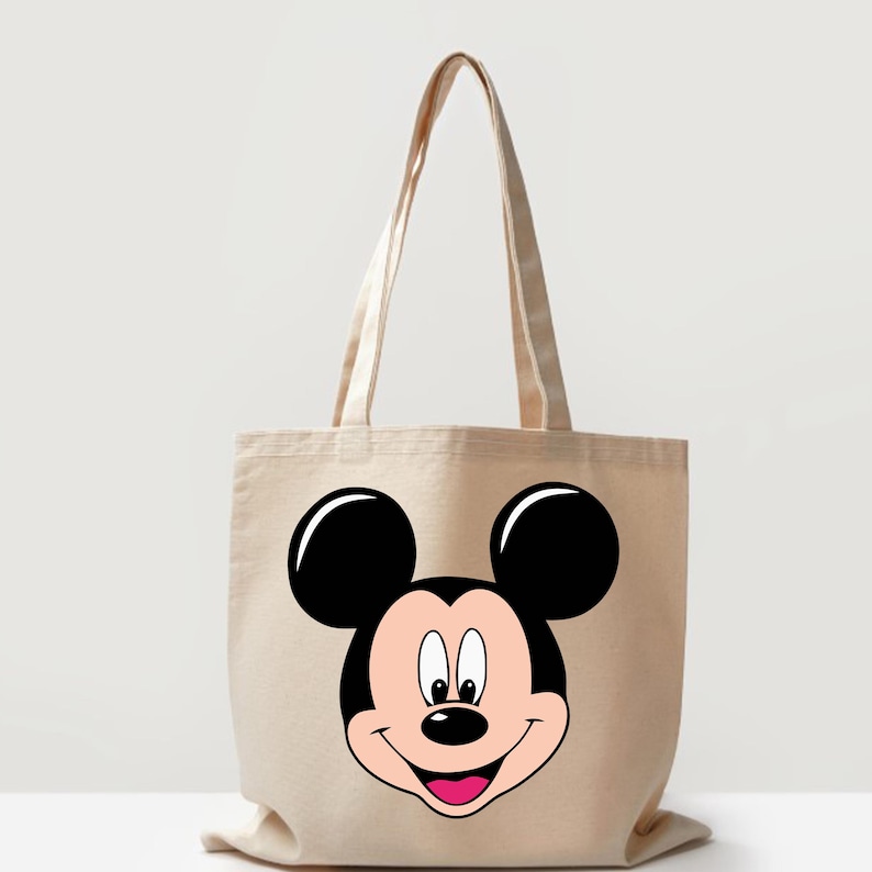 May include: Beige canvas tote bag featuring a large graphic of Mickey Mouse's face. The design includes black ears, a black nose, a pink mouth, and a light pink face. The bag has two beige handles.