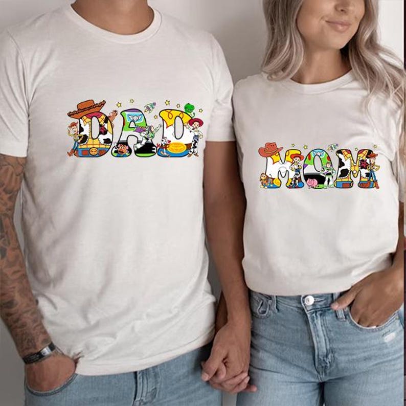 May include: Two white t-shirts with a Toy Story theme. The shirt on the left says "DAD" with the letters made up of Toy Story characters. The shirt on the right says "MOM" with the letters made up of Toy Story characters.