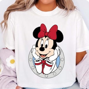 May include: White t-shirt featuring a cartoon Minnie Mouse design. Minnie Mouse is wearing a red bow, white gloves, and a red and white outfit. The character is framed by a gray circle with white accents.