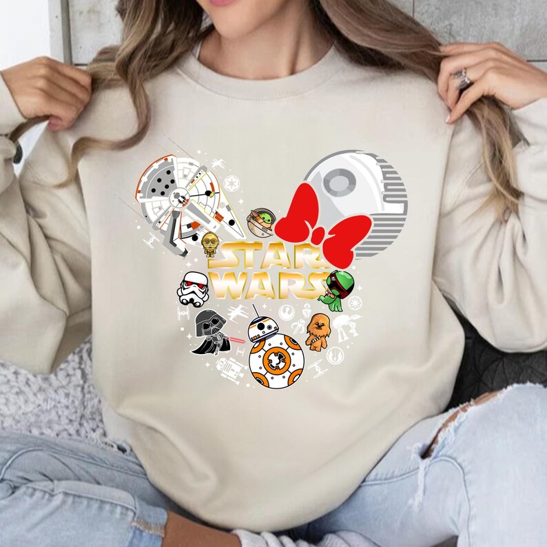 May include: A beige sweatshirt with a Star Wars themed design. The design features a Mickey Mouse head silhouette with Star Wars characters and a red bow. The text "STAR WARS" is written in gold letters.