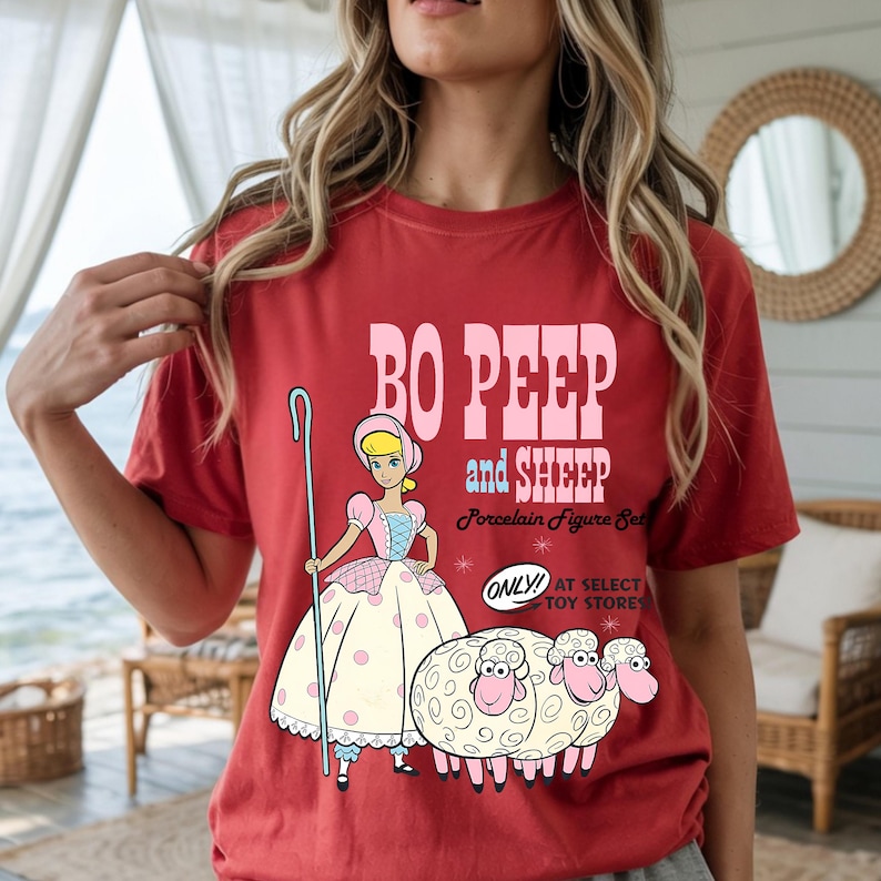 May include: Red t-shirt with a cartoon illustration of Bo Peep and two sheep, with the text "Bo Peep and Sheep Porcelain Figure Set." The design includes Bo Peep holding a shepherd's crook. The shirt has a vintage, distressed appearance.