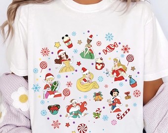 May include: White t-shirt featuring colorful Christmas-themed illustrations of Disney princesses. The design includes snowflakes, candy canes, stars, and festive beverages. The princesses are wearing Santa hats and holiday attire.