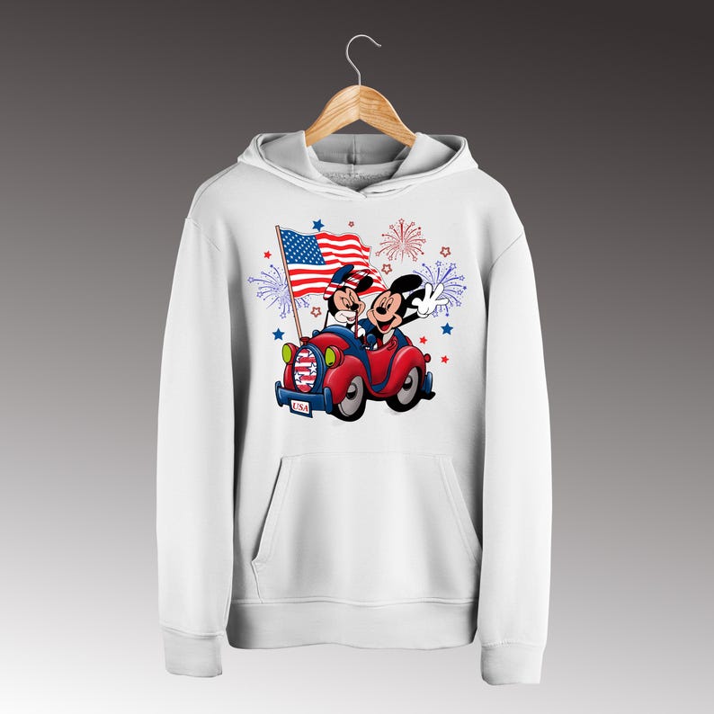 May include: White hooded sweatshirt featuring a graphic of Mickey Mouse and Minnie Mouse in a red car, waving an American flag. The design includes fireworks and stars, with the word "USA" on the car. A patriotic theme.