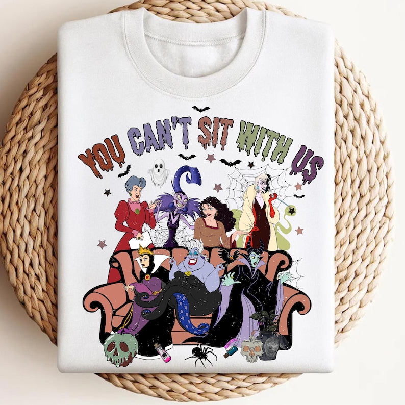 May include: White sweatshirt featuring a colorful graphic of Disney villains seated on a couch. The phrase "YOU CAN'T SIT WITH US" is printed above the characters. The design includes bats, stars, and a spiderweb.