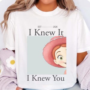 May include: A white short-sleeved t-shirt featuring a graphic print of a cartoon character wearing a pink hat. The text "EST 2026", "I Knew It", and "I Knew You" is printed in black lettering above and below the central square illustration.