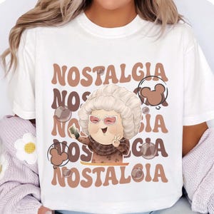 May include: White t-shirt with the word "NOSTALGIA" in brown retro font. The shirt features a cartoon of a smiling elderly person wearing sunglasses and a scarf, holding a phone and making a peace sign.