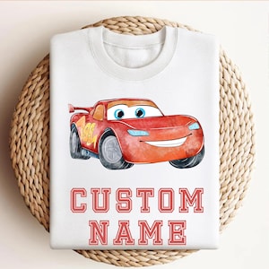 May include: White sweatshirt featuring a red cartoon race car with the number 95. The car has blue eyes and a smiling mouth. Below the car, the text "CUSTOM NAME" is printed in red block letters.