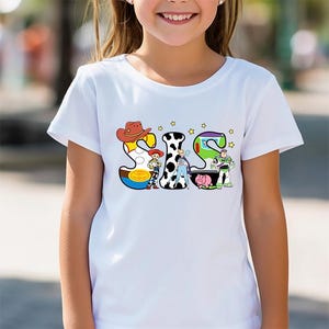 May include: White t-shirt with a colorful graphic of the word "SIS" made up of characters from the Toy Story movie franchise. The characters include Woody, Jessie, Buzz Lightyear, and Rex.
