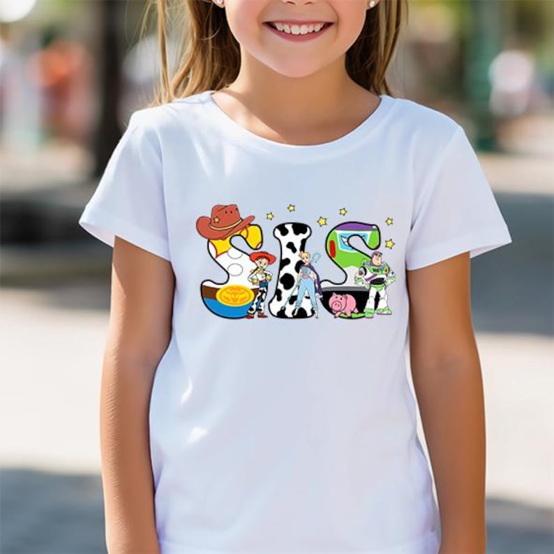 May include: White t-shirt with a colorful graphic of the word "SIS" made up of characters from the Toy Story movie franchise. The characters include Woody, Jessie, Buzz Lightyear, and Rex.