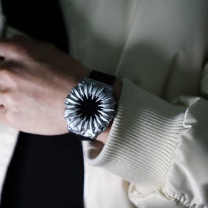 May include: A silver watch with a unique, circular, flower-like design. The watch face is black and the band is black leather.