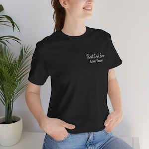 Op de afbeelding: Zwart T-shirt met de tekst "Best Dad Ever, Love, Emma" in witte letters. Het shirt heeft een klassieke ronde hals en lijkt gemaakt van een zacht materiaal. De tekst staat linksboven.