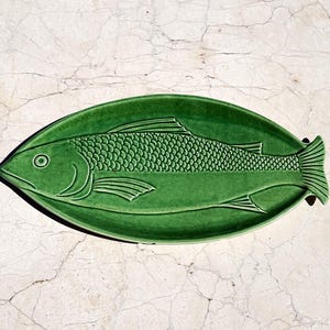 Puede incluir: Una bandeja de servicio ovalada de cerámica con un esmalte verde esmeralda intenso. El plato presenta un diseño en relieve de un pez que abarca toda la longitud de la bandeja, mostrando escamas detalladas, aletas y un ojo definido.