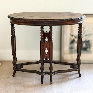 May include: An oval wooden side table with a dark brown finish, featuring a tabletop with a central diamond inlay pattern. The table is supported by four turned legs connected by a central vertical support and a curved, open-work base.