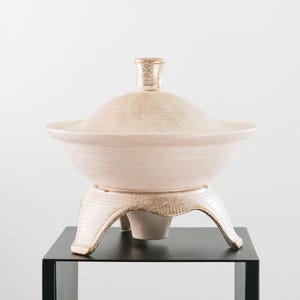 May include: A light pink ceramic serving bowl with a lid and a decorative handle. The bowl sits on a matching stand with three curved legs. The bowl and lid have a textured surface.