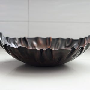 Puede incluir: Un cuenco decorativo de metal oscuro con un borde ondulado. El cuenco tiene un acabado met&aacute;lico con tonos marrones y negros. El cuenco est&aacute; sobre una superficie blanca.