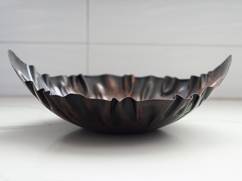 Puede incluir: Un cuenco decorativo de metal oscuro con un borde ondulado. El cuenco tiene un acabado met&aacute;lico con tonos marrones y negros. El cuenco est&aacute; sobre una superficie blanca.