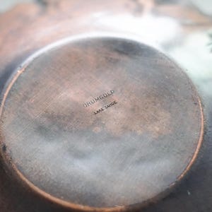 Puede incluir: Primer plano del interior de un cuenco de metal color cobre. La base presenta una zona circular ligeramente elevada con el texto estampado "DRUMGOLD LAKE TAHOE". La superficie del cuenco tiene un aspecto texturizado y envejecido, con variaciones de color.
