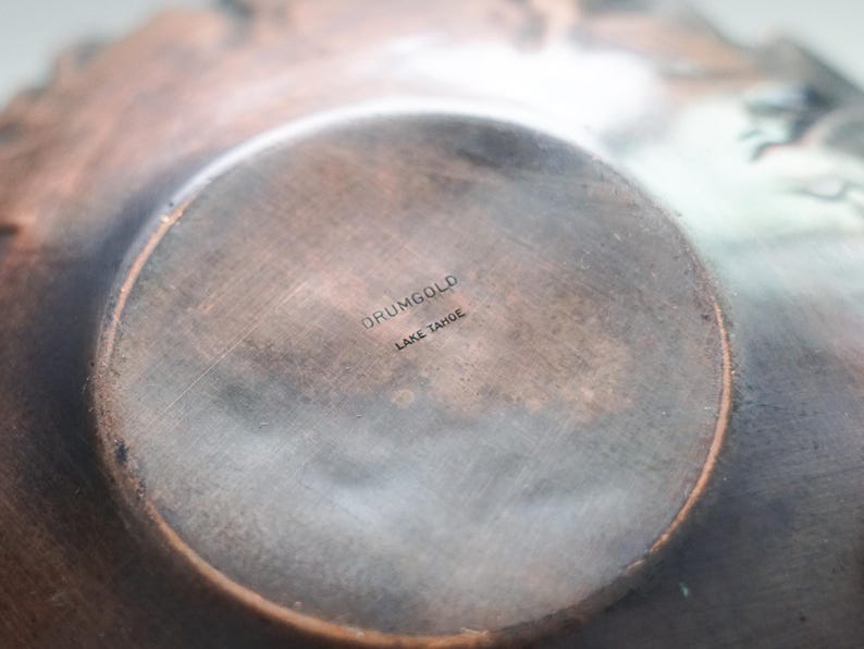 Puede incluir: Primer plano del interior de un cuenco de metal color cobre. La base presenta una zona circular ligeramente elevada con el texto estampado "DRUMGOLD LAKE TAHOE". La superficie del cuenco tiene un aspecto texturizado y envejecido, con variaciones de color.