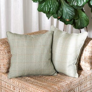 Peut inclure: Deux coussins carrés de couleur vert sauge clair avec un motif à carreaux subtil composé de fines lignes marron clair. Chaque coussin présente une finition passepoilée nette et un aspect de tissu texturé, formant une paire assortie.
