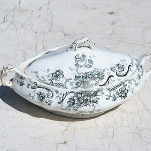 May include: A white ceramic tureen with a lid, featuring a detailed black floral and scrollwork pattern. The oval-shaped serving dish includes two side handles and a central lid handle, all accented with subtle gold trim along the edges.