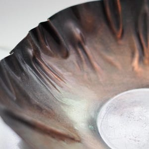 Puede incluir: Primer plano de un cuenco artesanal con tonos cobrizos y borde ondulado. La superficie del cuenco tiene un brillo met&aacute;lico con tonos marrones y plateados. El dise&ntilde;o presenta un peque&ntilde;o elemento redondo plateado.