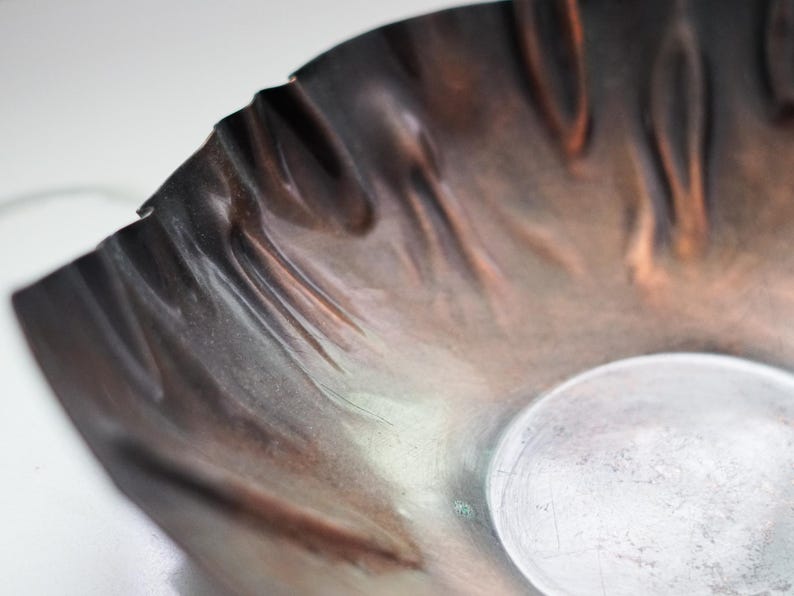 Puede incluir: Primer plano de un cuenco artesanal con tonos cobrizos y borde ondulado. La superficie del cuenco tiene un brillo met&aacute;lico con tonos marrones y plateados. El dise&ntilde;o presenta un peque&ntilde;o elemento redondo plateado.