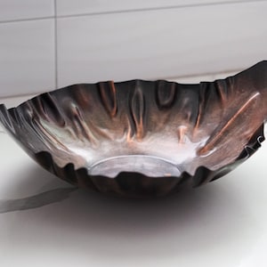 Puede incluir: Un cuenco decorativo de metal hecho a mano con un borde &uacute;nico y ondulado. El cuenco tiene un acabado oscuro y bru&ntilde;ido con toques de cobre y bronce, creando una apariencia texturizada y org&aacute;nica. El cuenco est&aacute; dise&ntilde;ado para ser una pieza central o un art&iacute;culo decorativo.