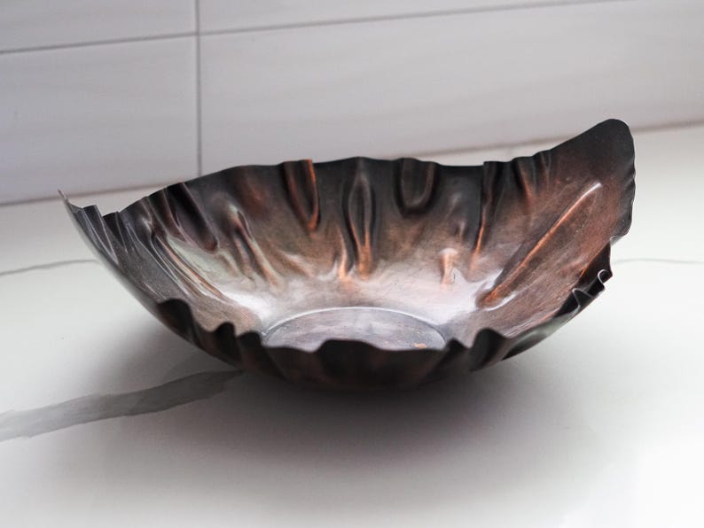 Puede incluir: Un cuenco decorativo de metal hecho a mano con un borde &uacute;nico y ondulado. El cuenco tiene un acabado oscuro y bru&ntilde;ido con toques de cobre y bronce, creando una apariencia texturizada y org&aacute;nica. El cuenco est&aacute; dise&ntilde;ado para ser una pieza central o un art&iacute;culo decorativo.