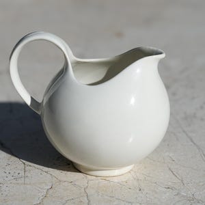 May include: A white ceramic creamer with a rounded body and a curved handle. The creamer has a small spout for pouring and sits on a circular base. The design is simple and elegant, suitable for serving cream or milk.