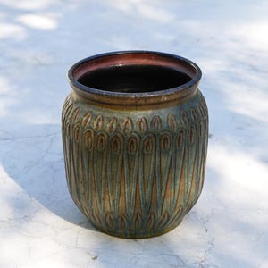 May include: A ceramic pot featuring a dark brown and teal glaze with a repeating vertical teardrop pattern embossed around the exterior. The rim is smooth and dark, while the interior is deep and dark, showcasing a rounded, wide-mouthed shape.