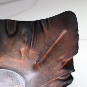 Puede incluir: Primer plano de un cuenco de metal texturizado de color cobre con una forma org&aacute;nica &uacute;nica. El cuenco tiene un acabado oscuro y bru&ntilde;ido con variaciones visibles en la superficie y un interior m&aacute;s claro, plateado.