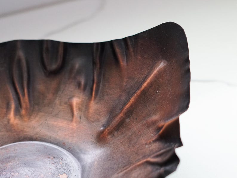 Puede incluir: Primer plano de un cuenco de metal texturizado de color cobre con una forma org&aacute;nica &uacute;nica. El cuenco tiene un acabado oscuro y bru&ntilde;ido con variaciones visibles en la superficie y un interior m&aacute;s claro, plateado.