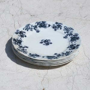 Puede incluir: Una pila de cuatro platos redondos de cerámica blanca con un patrón floral azul oscuro a lo largo de los bordes festoneados y un pequeño grupo floral a juego en el centro de cada plato. Los platos tienen un acabado liso y uniforme.