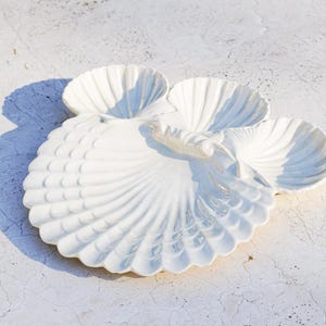 Könnte beinhalten: Weißes Serviertablett aus Keramik in Muschelform mit vier Fächern. Das Tablett hat eine große, fächerförmige Basis mit einem gewellten Rand und drei kleinere muschelförmige Fächer. Auf einer weißen, strukturierten Oberfläche.