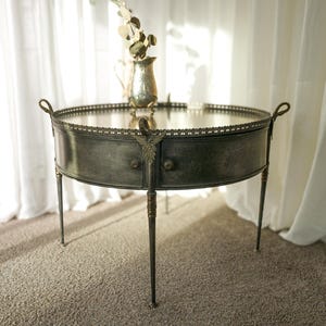 May include: An antique silver-toned oval side table with a mirrored top and decorative trim. The table has curved sides, two small drawers, and slender legs. A small vase with decorative branches sits on top.