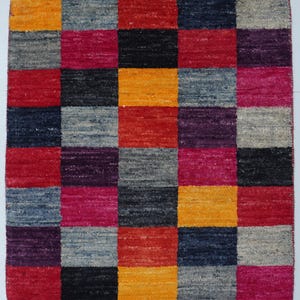 Könnte beinhalten: Ein quadratischer, mehrfarbiger Teppich mit Schachbrettmuster. Der Teppich hat Quadrate in Rot, Orange, Schwarz, Grau, Lila und Pink. Der Teppich besteht aus einem gewebten Material.