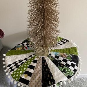 May include: A white, black, and green patchwork Christmas tree skirt with a white pom-pom trim. A gold artificial Christmas tree sits on top of the skirt.