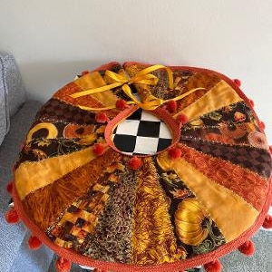 May include: A handmade autumn-themed tree skirt with a patchwork design featuring various fall colors and patterns. The skirt is decorated with a red pom-pom trim and yellow ribbon ties.