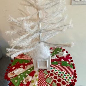 May include: A white artificial Christmas tree with a colorful tree skirt. The skirt is a patchwork design with red, green, and white fabrics, accented with red pom-poms. The tree is set on a black and white striped stand.