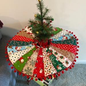 May include: A colorful patchwork Christmas tree skirt with red pom-poms around the edge. A small green Christmas tree with gold ornaments sits on top of the skirt.