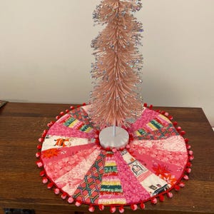 May include: A pink bottle brush Christmas tree sits on a circular tree skirt. The tree skirt is made of pink fabric with a variety of patterns and a red pom-pom trim. The words "Merry & Bright" are printed on the skirt.