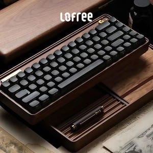 Puede incluir: Un teclado mecánico negro con teclas redondas se encuentra en una caja de madera. El teclado tiene un esquema de color negro y marrón y el nombre de la marca "LOFREE" está impreso en la caja.