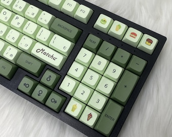 May include: A green and black keyboard with keycaps featuring various food and drink icons, including ice cream, matcha, and a hamburger. The keyboard has a 'Matcha' keycap and a 'NUM' keycap.
