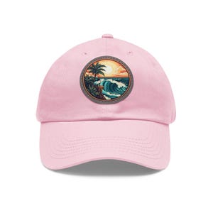 Op de afbeelding: Roze baseballpet met een ronde patch met een zonsondergangscène met een palmboom, een surfer en een golf.