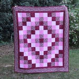 May include: A rectangular quilt featuring a patchwork design of alternating light pink, dark magenta, and floral-patterned fabric squares. The quilt is finished with a wide floral-patterned border and displays intricate circular machine quilting.