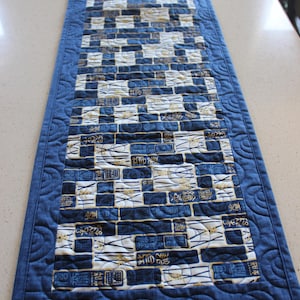May include: A rectangular table runner with a brick-like pattern in shades of blue, white, and gold. The runner has a solid blue border with a quilted, curved design. The fabric features Asian-inspired symbols.