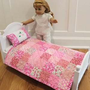 May include: A white wooden doll bed with a pink patchwork quilt and pillow. The quilt features various floral patterns in shades of pink. The pillow has a floral embroidery design. The bed is designed for dolls.