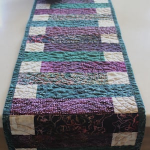 May include: A quilted table runner with a patchwork design. The runner features alternating sections of teal, purple, and white fabric. The edges are finished with a teal border. A gray bowl with a unique shape sits on the runner.